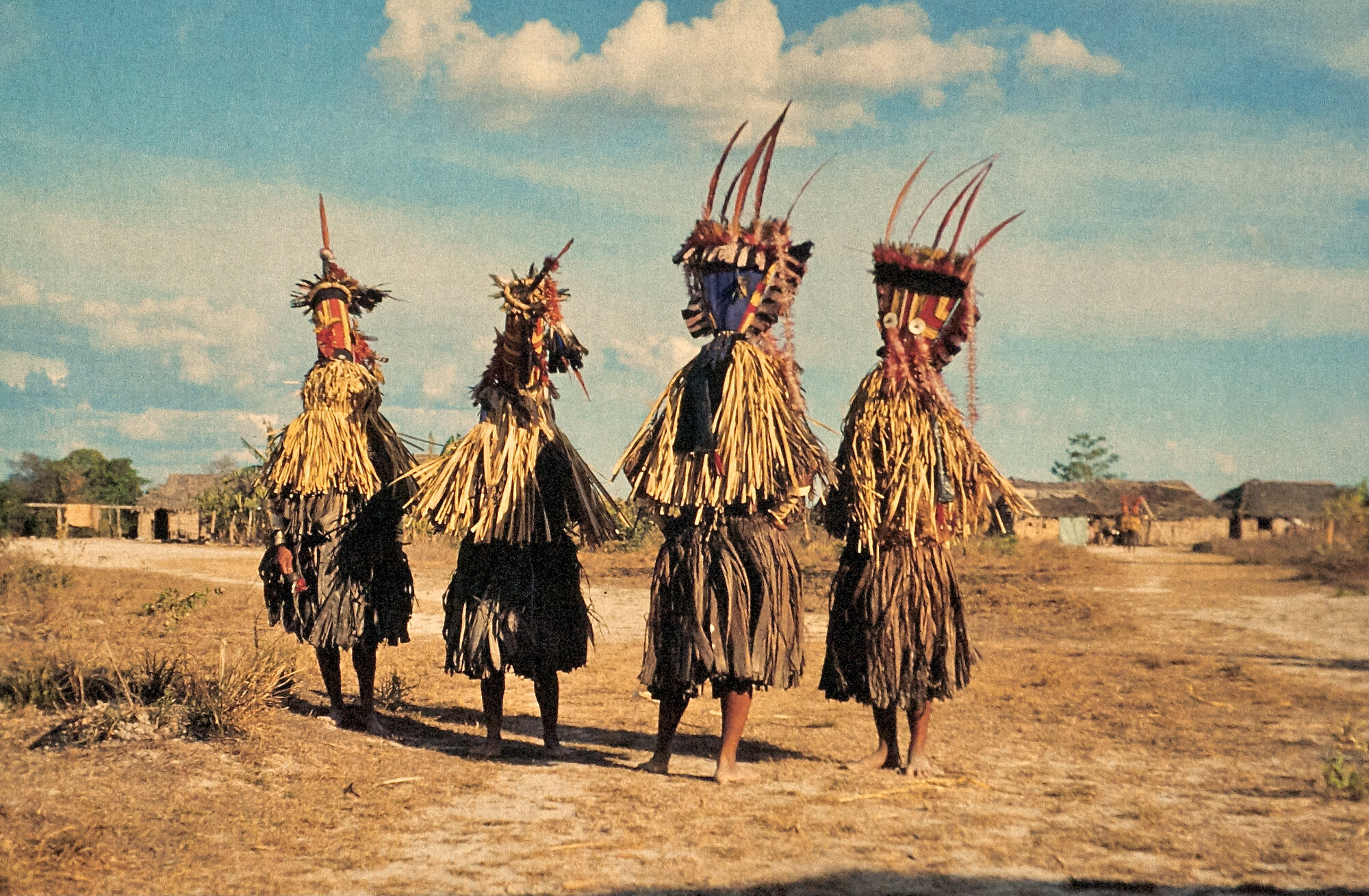 Three figures in elaborate ceremonial straw costumes and painted masks standing in an open field