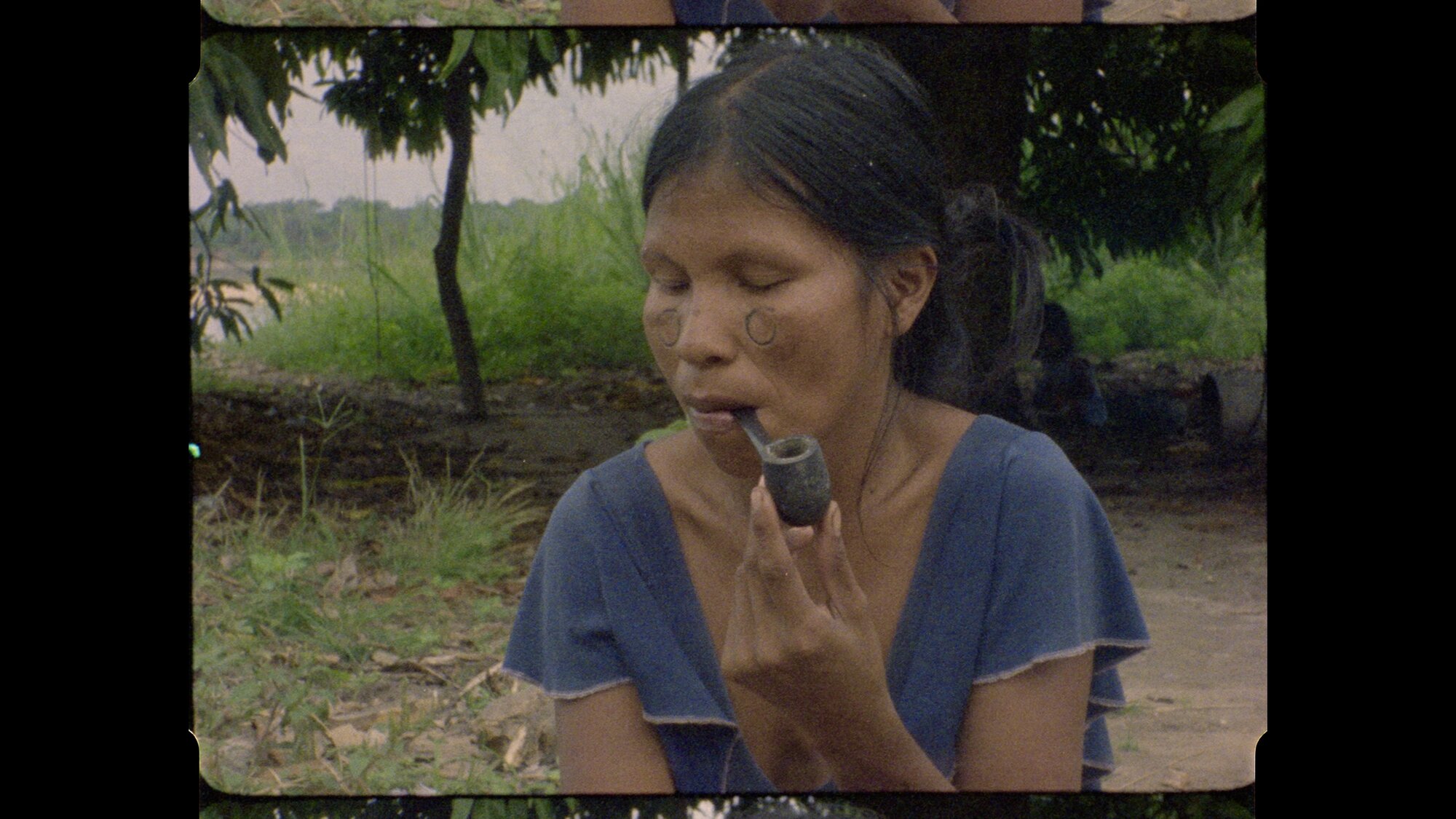 Diriti wearing a blue top with traditional face paint, painting a small clay doll outdoors