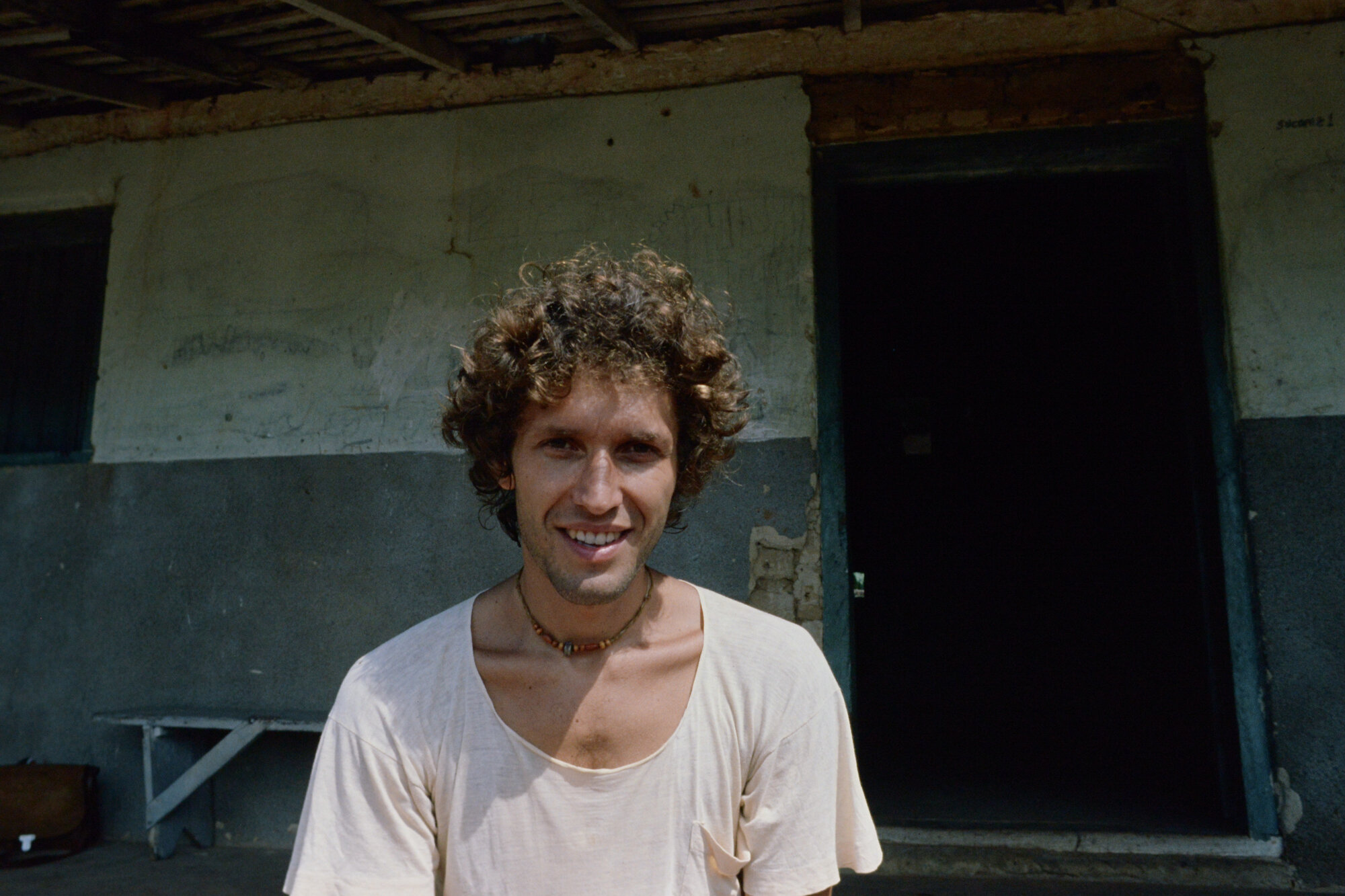 Young Matti with curly hair and a beaded necklace, smiling in front of a village building