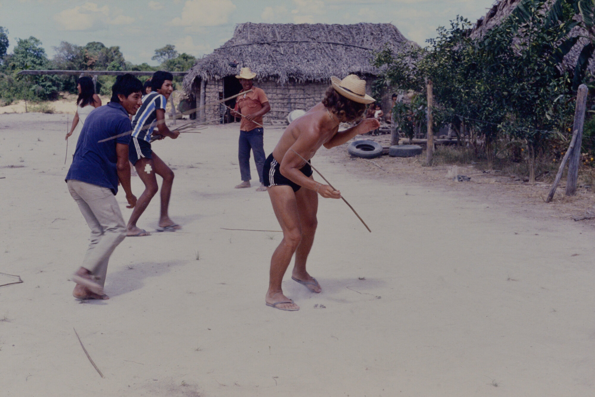 Matti playing a game with indigenous villagers in a sandy area with thatched huts in the background
